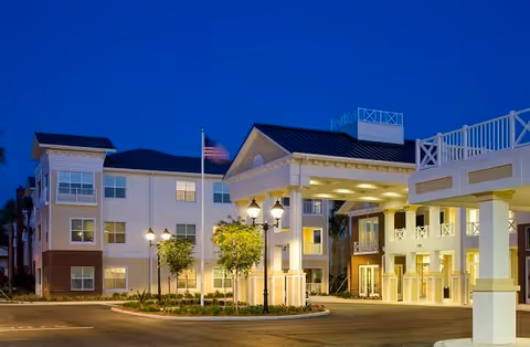 Exterior view of Watercrest Buena Vista senior living facility at dusk, showing a well-lit entrance with a covered drop-off area, multiple windows, and an American flag on a flagpole. The building has three stories with white and beige walls and decorative railings on the balconies.