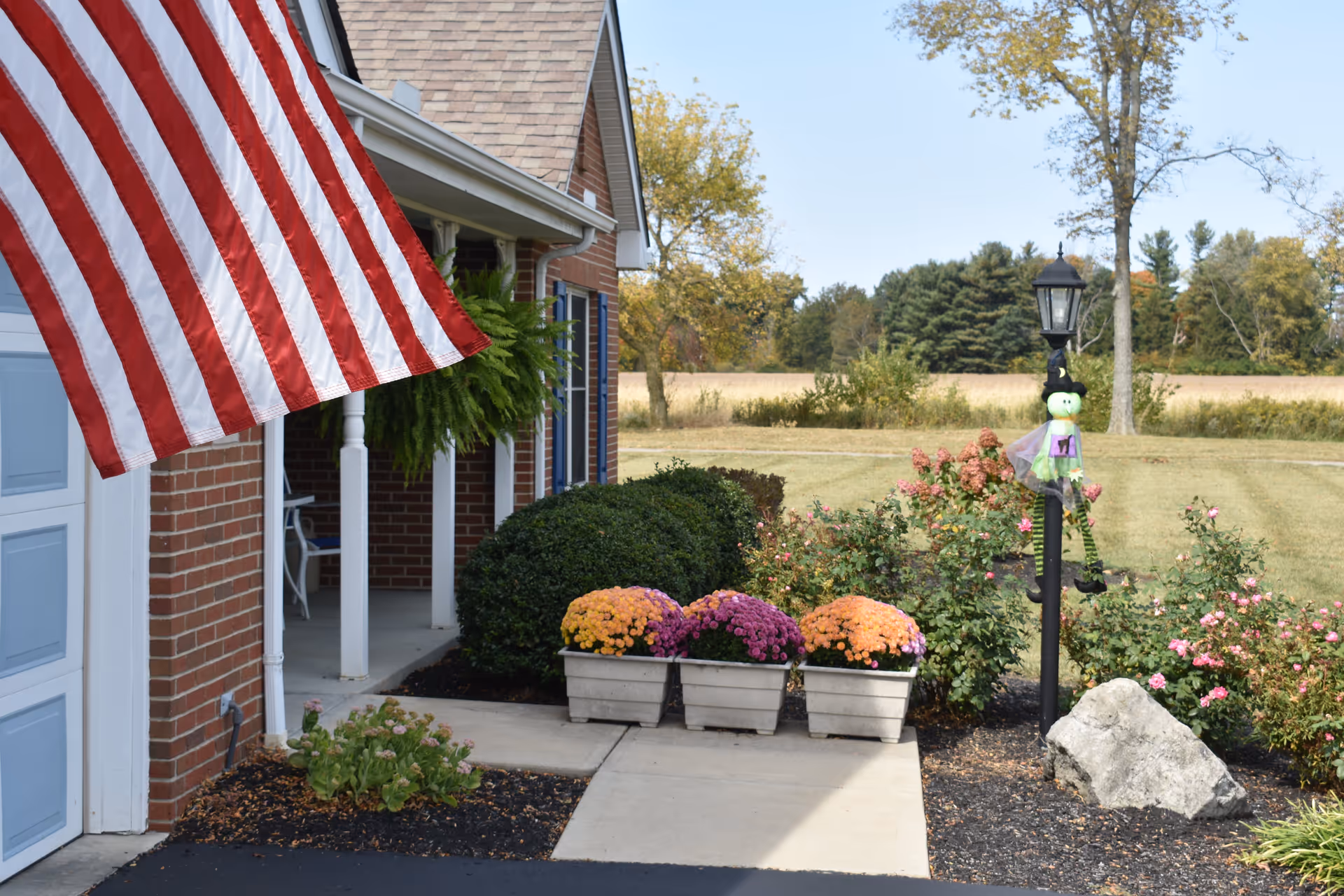 Front exterior view of a brick building with a large American flag hanging near the garage. There is a porch with white columns and a hanging green plant. In front of the porch, there are three planters with colorful flowers and a black lamp post decorated with a small green witch figure. The background shows a grassy field and trees under a clear sky.