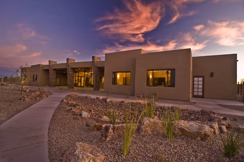 Modern single-story beige building with large windows, desert landscaping, and a paved walkway.