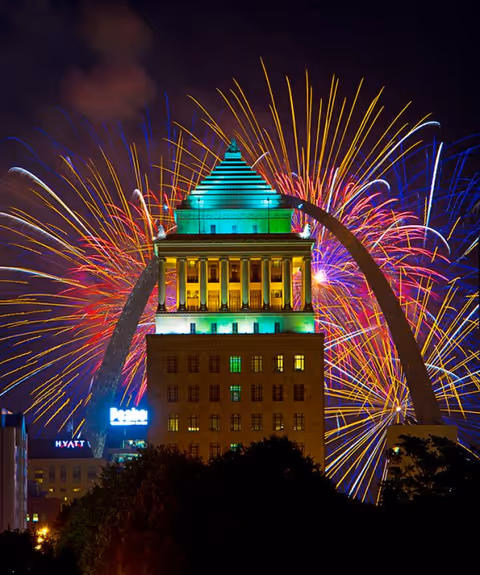 Nighttime cityscape with a lit, multi-tiered building surrounded by colorful fireworks.