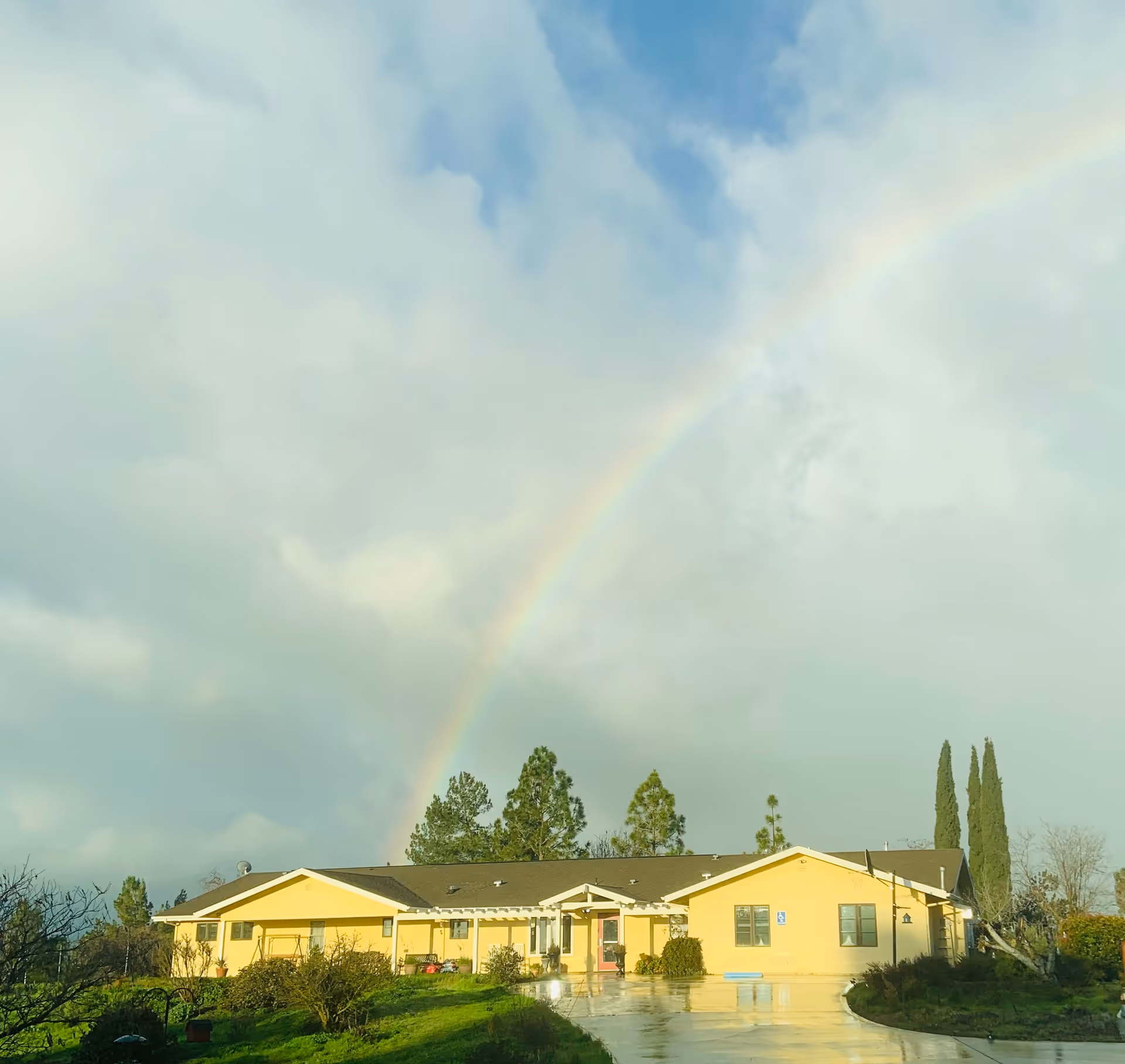 Yellow assisted living building sits beneath a rainbow in a cloudy sky.