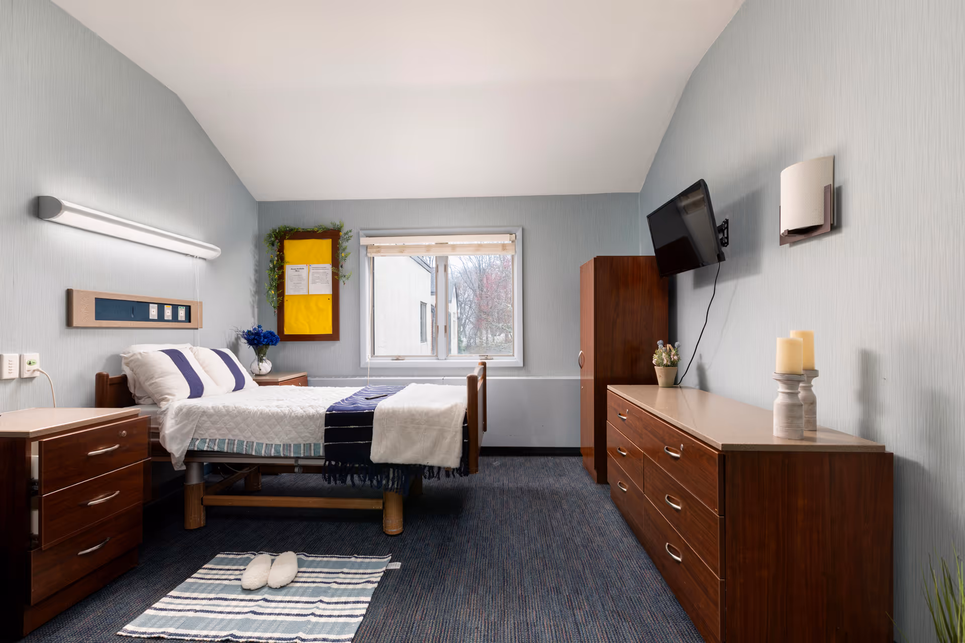 A furnished senior living bedroom with a bed, dresser, wall-mounted TV, and window.