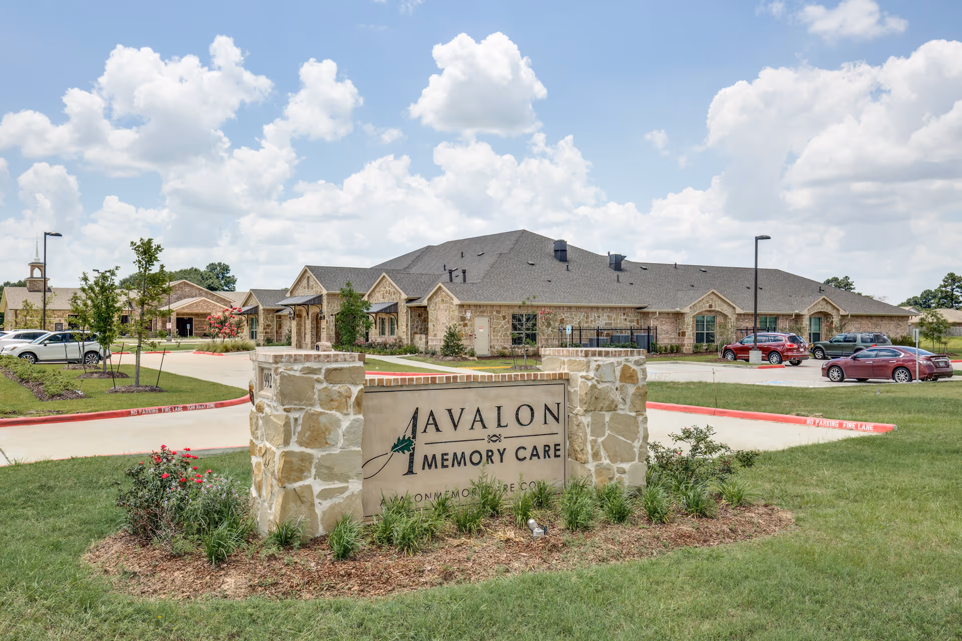 Exterior view of Avalon Memory Care facility showing a stone sign with the facility name in front of a large building with a parking lot and several cars under a partly cloudy sky.
