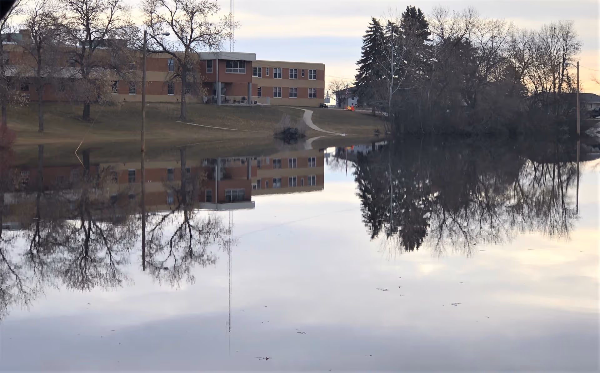A low brick medical building on a grassy slope reflected in a calm pond with leafless trees.