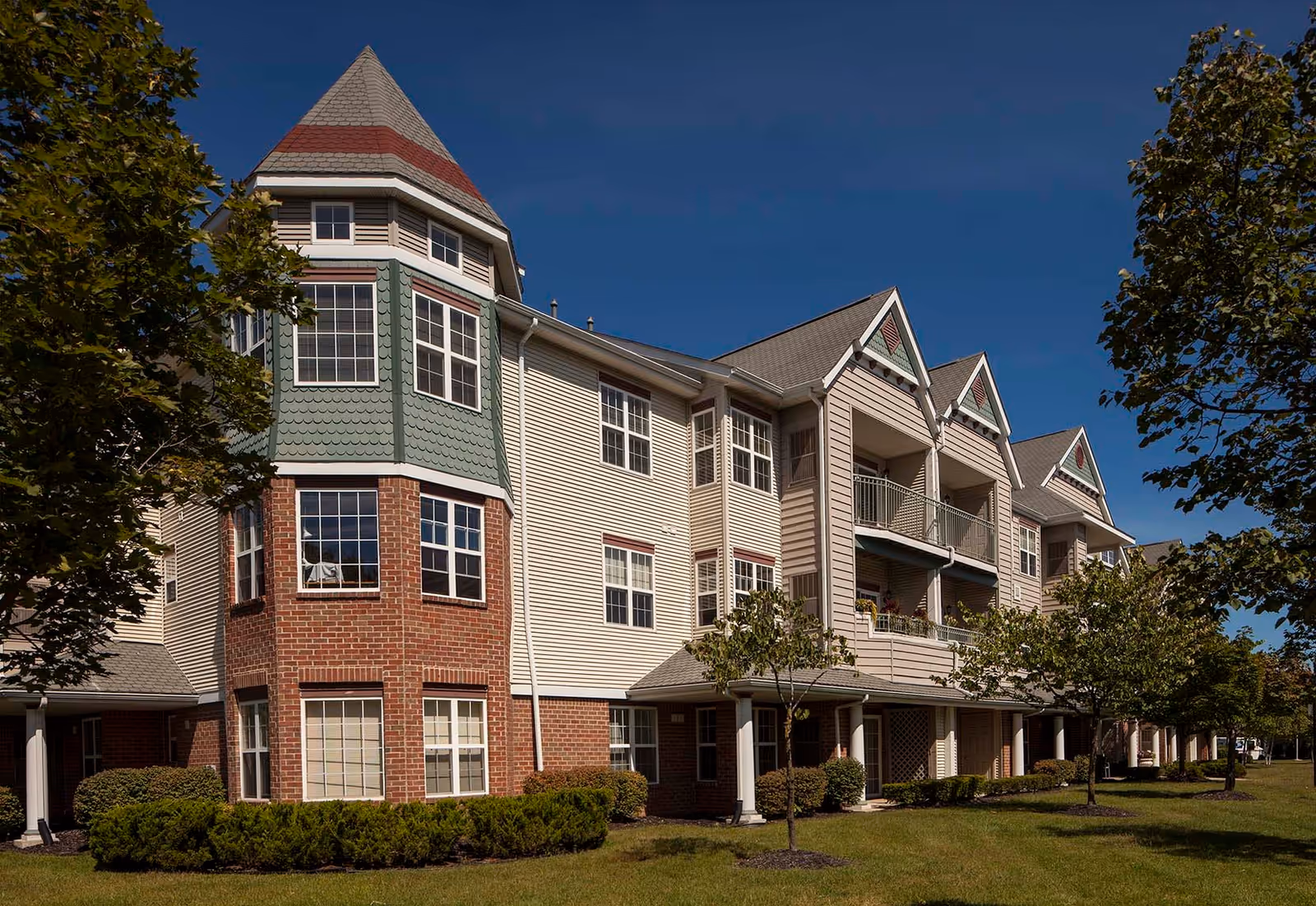 Exterior view of a multi-story senior living facility building with a combination of brick and siding, featuring a turret-like structure on the left and balconies on the right, surrounded by green lawns and trees under a clear blue sky.