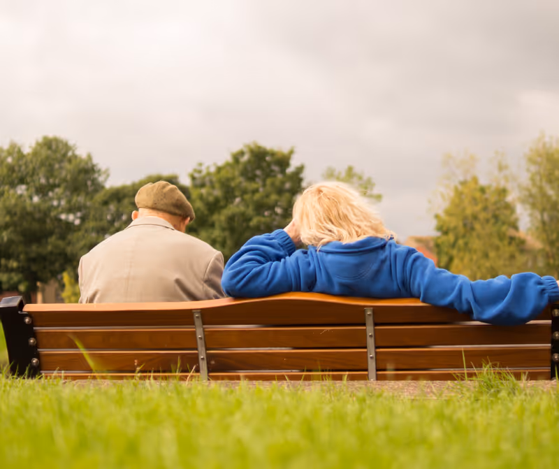 Two seniors sit on a wooden bench in a park, facing away.