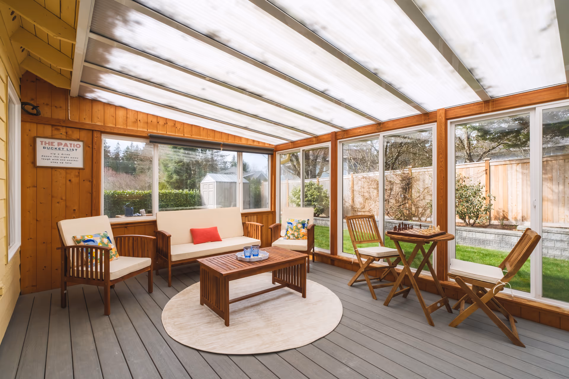 Enclosed glass-walled patio with wooden seating, a coffee table, and a round rug.