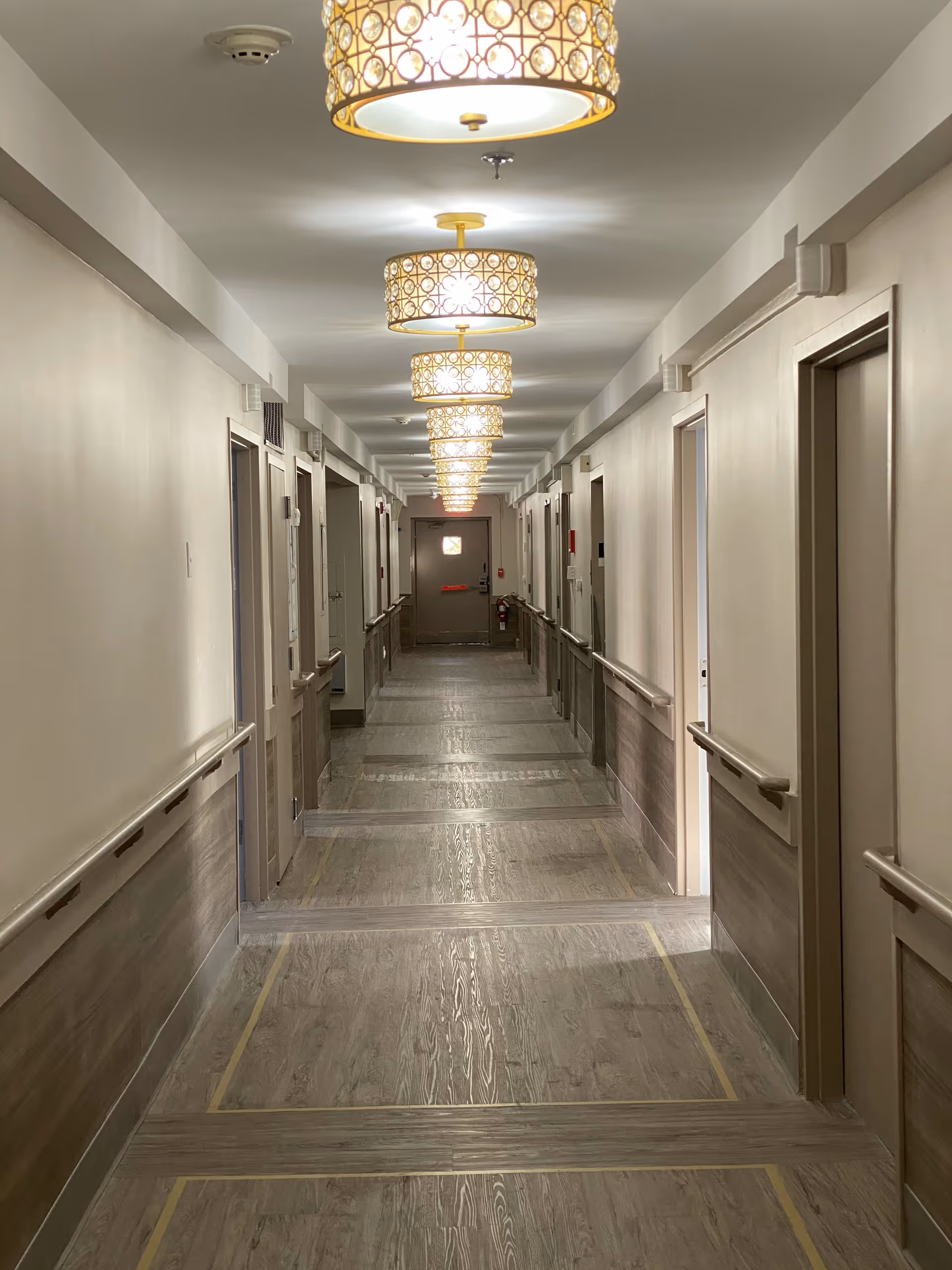 A long, well-lit hallway in a senior living or rehabilitation facility with multiple closed doors on both sides, handrails along the walls, and decorative ceiling lights.