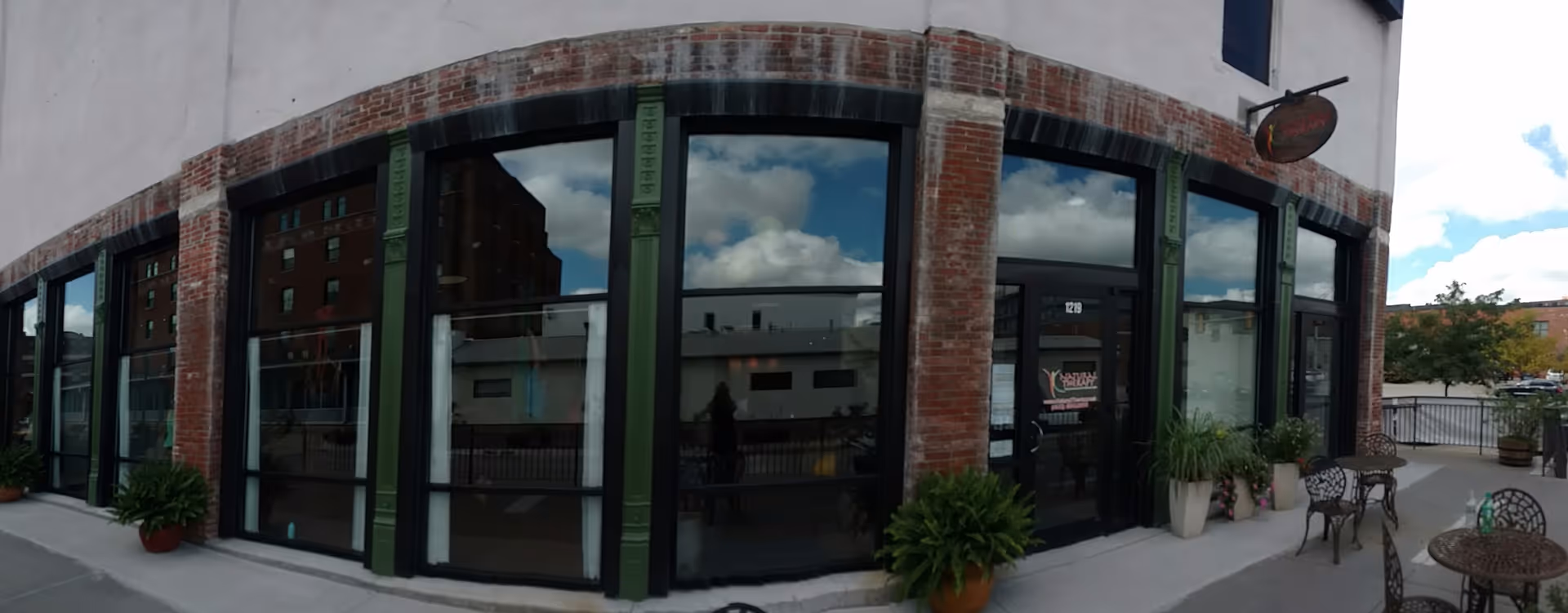 Exterior brick storefront with large glass windows, potted plants, and outdoor seating.