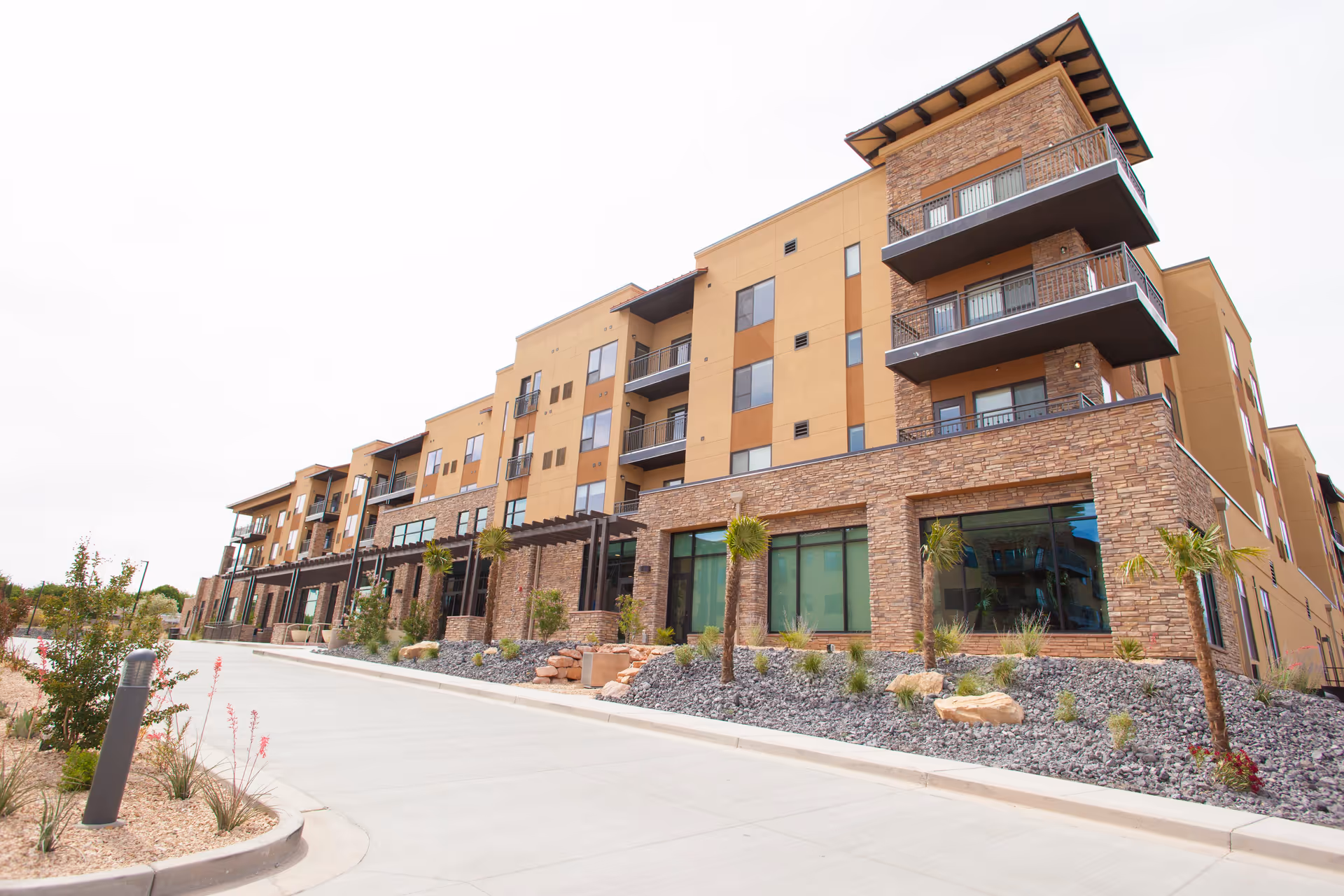 Exterior view of a modern multi-story building with stone and beige stucco facade, balconies, large windows, and landscaped surroundings including small palm trees and shrubs along a paved driveway.