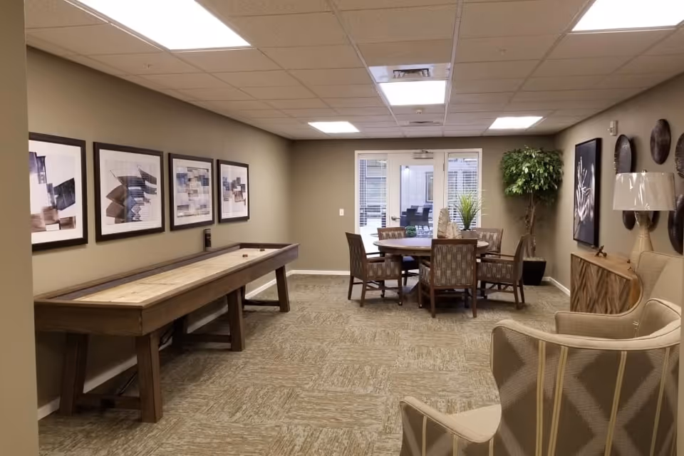 Interior common lounge with a shuffleboard table along the left wall, a round table and chairs near the back, and upholstered armchairs in the foreground.