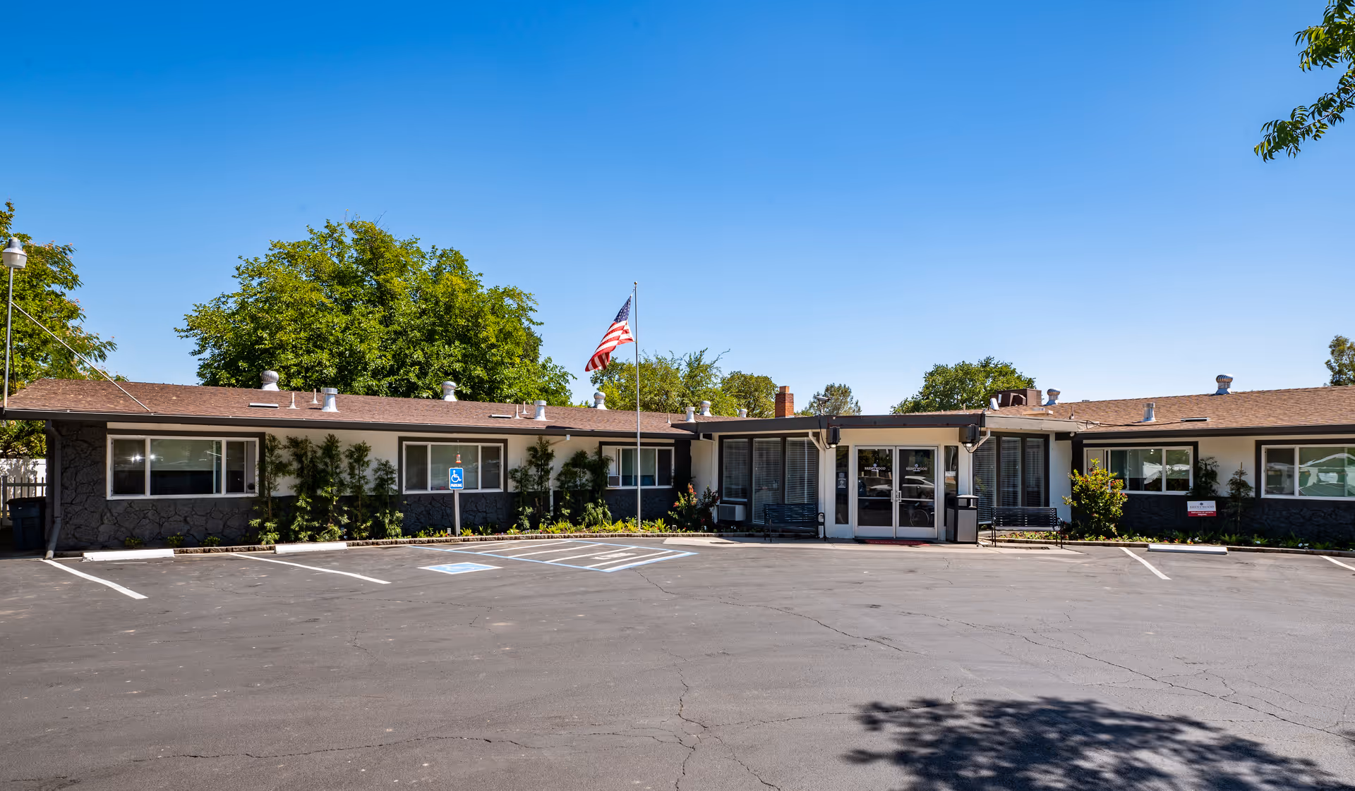 Front view of a single-story senior living facility with an American flag, entrance doors, and an empty parking lot under a clear blue sky.