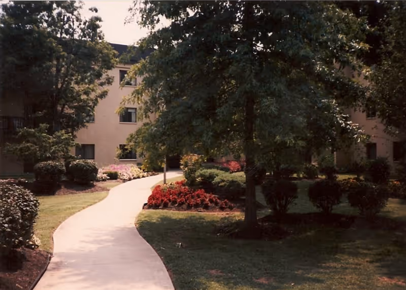 Curved concrete walkway through landscaped lawns and flowerbeds leading to the entrance of a multi-story residence framed by trees.