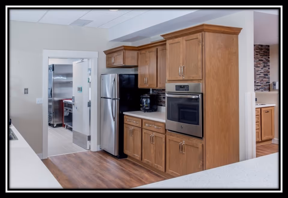 Interior view of a kitchen area with wooden cabinets, a stainless steel refrigerator, and a built-in oven. The floor is wooden, and there is a doorway leading to another room with stainless steel kitchen equipment visible.