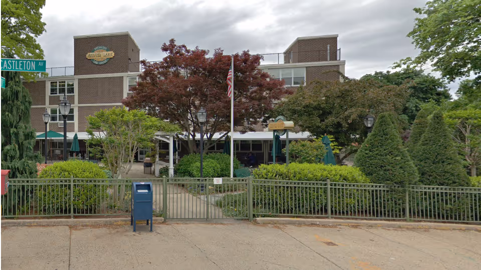 Front exterior view of Silver Lake Specialized Care Center, a multi-story brick building with a sign displaying the facility name. The entrance is surrounded by well-maintained greenery, trees, and bushes, with a metal fence and gate in front. A blue mailbox is positioned on the sidewalk outside the fence, and there is a street sign for Castleton Avenue on the left side.