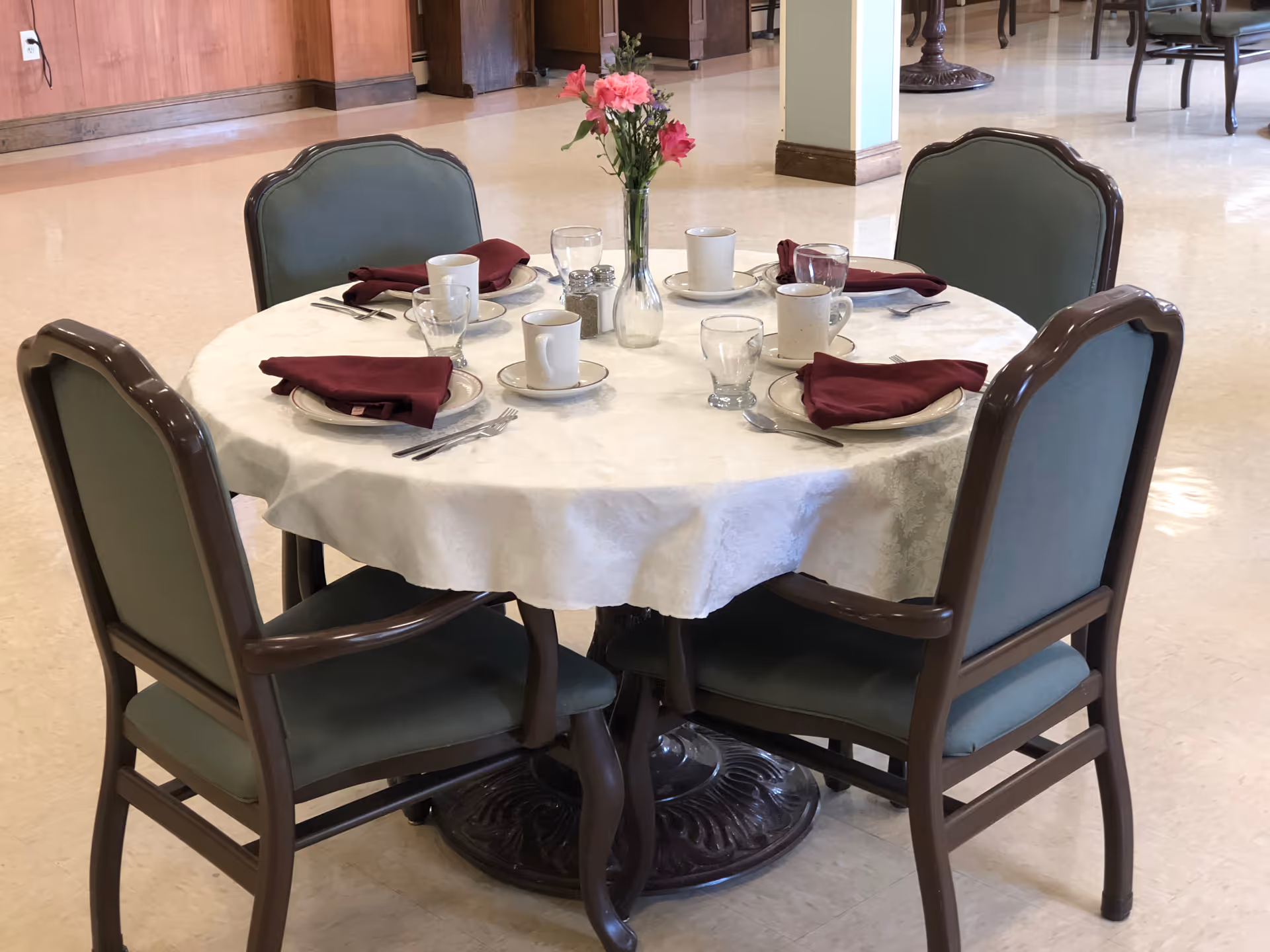 Round dining table in a communal dining area set with cups, glasses, silverware, folded burgundy napkins and a small vase of flowers.