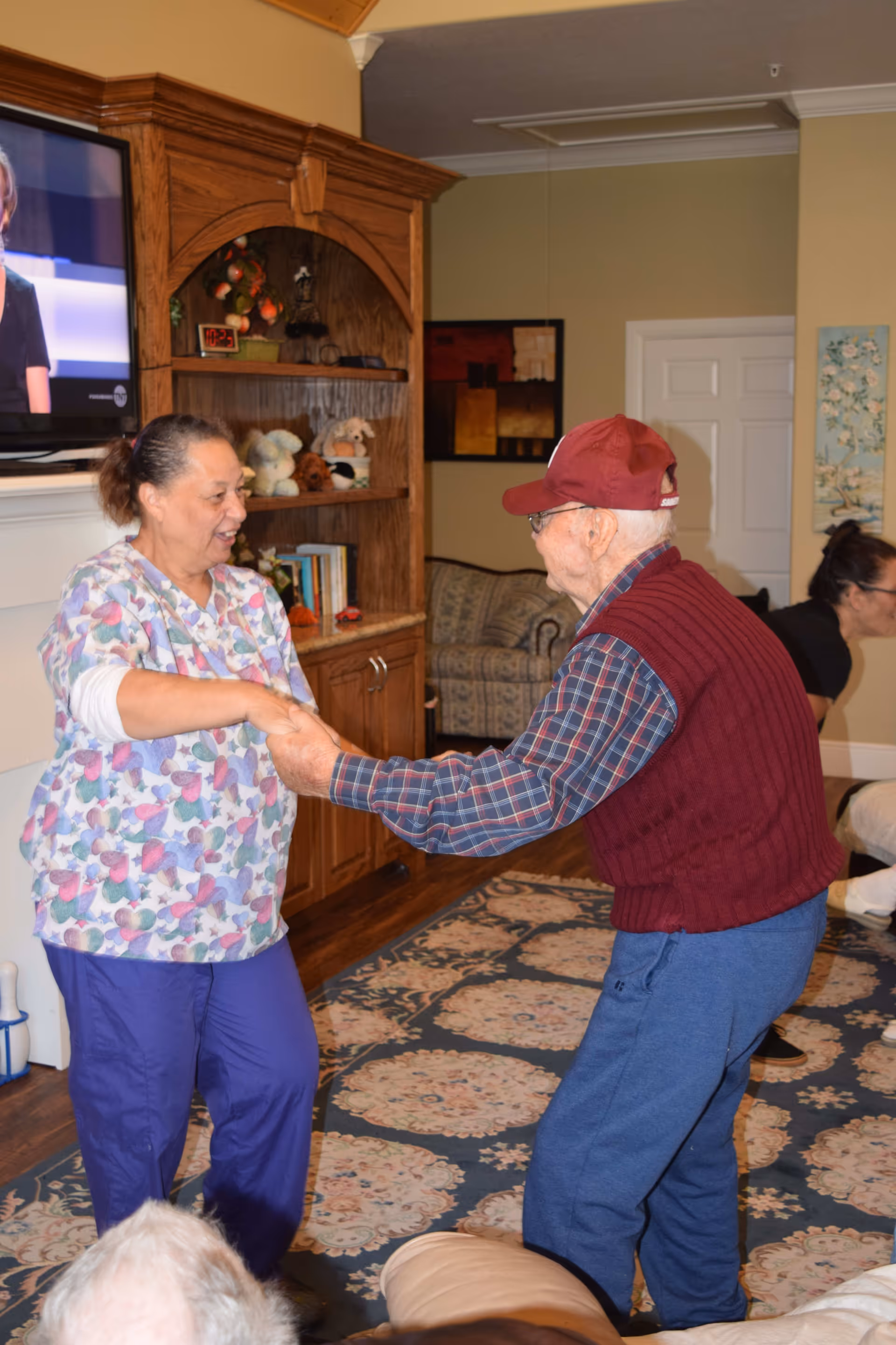 An elderly man wearing a red cap, plaid shirt, and maroon vest is holding hands and dancing with a woman in colorful scrubs in a cozy living room. The room has a patterned rug, wooden furniture with shelves holding stuffed animals and books, a TV mounted on the wall, and a couch in the background.