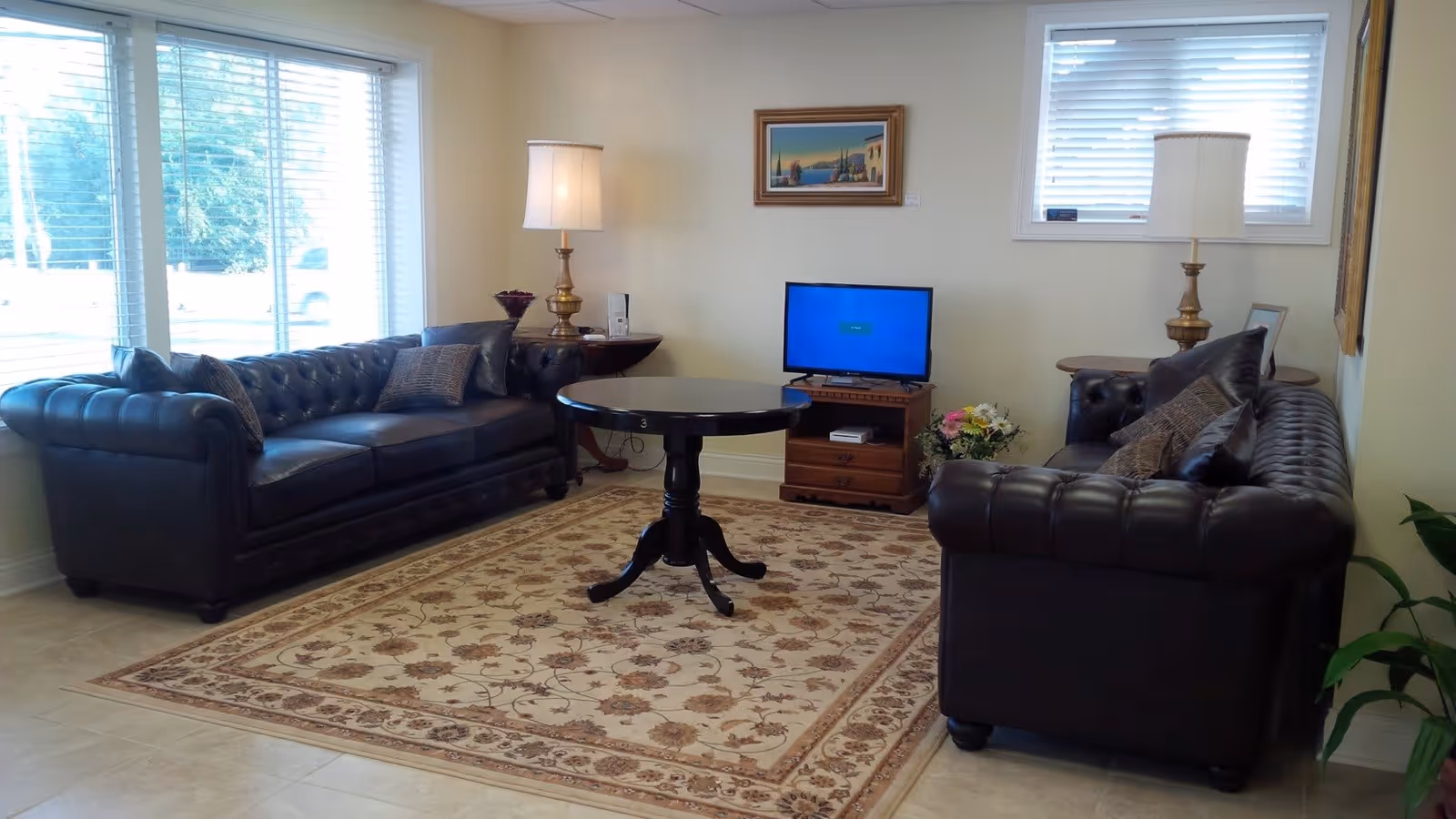 A cozy living room with two dark leather tufted sofas facing each other, a round black wooden table in the center on a floral patterned area rug. There are two brass table lamps with white shades on side tables behind each sofa, a small wooden TV stand with a flat-screen TV, a framed painting on the wall, and windows with white blinds letting in natural light.