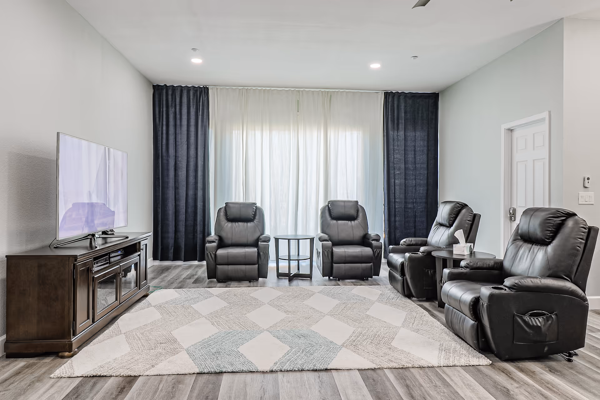 A bright living room with four black leather recliner chairs arranged around a large patterned area rug. A wooden TV stand with a flat-screen television is on the left side. The back wall features large windows covered with sheer white curtains and dark blue drapes. The floor is a light wood laminate.
