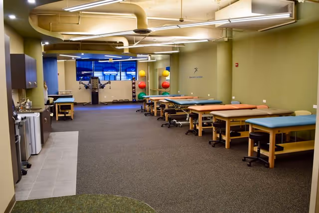 Long therapy room with treatment tables, exercise equipment, and mats in a rehab facility.