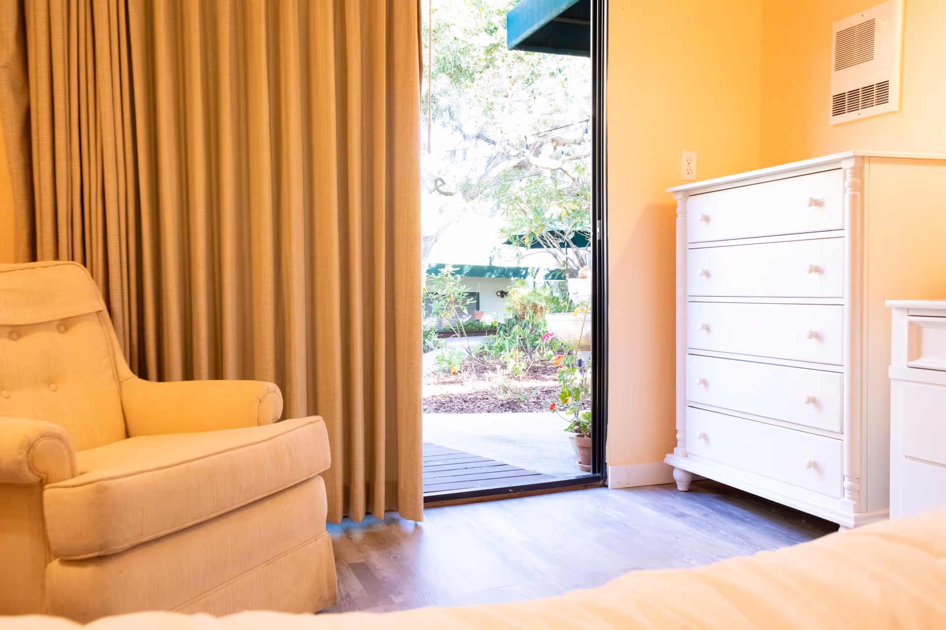 Cozy bedroom with beige armchair, white dresser, and sliding door opening to sunny outdoor patio.