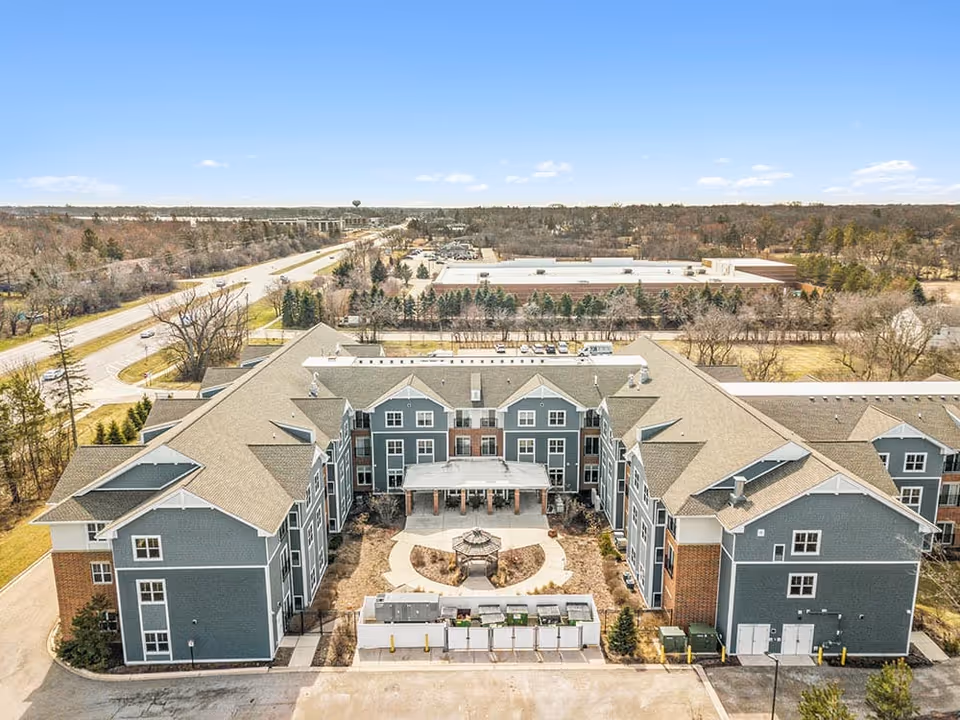 Aerial view of a large senior living facility building with a U-shaped layout, featuring a central courtyard with a gazebo and landscaped pathways. The building has gray siding with white trim and a beige roof. Surrounding the facility are trees, roads, and other buildings under a clear blue sky.