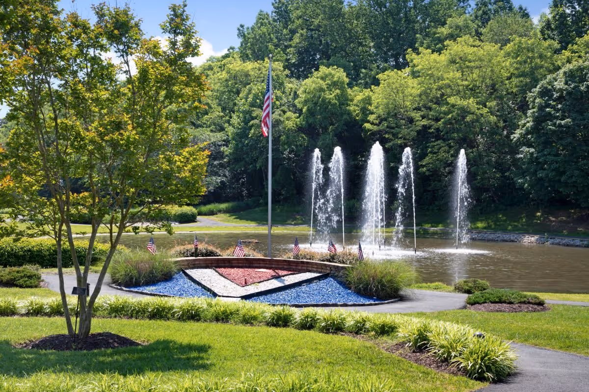 A landscaped outdoor scene featuring a flagpole, a red-white-and-blue decorative rock bed with small American flags, and multiple water fountains in a pond surrounded by trees.