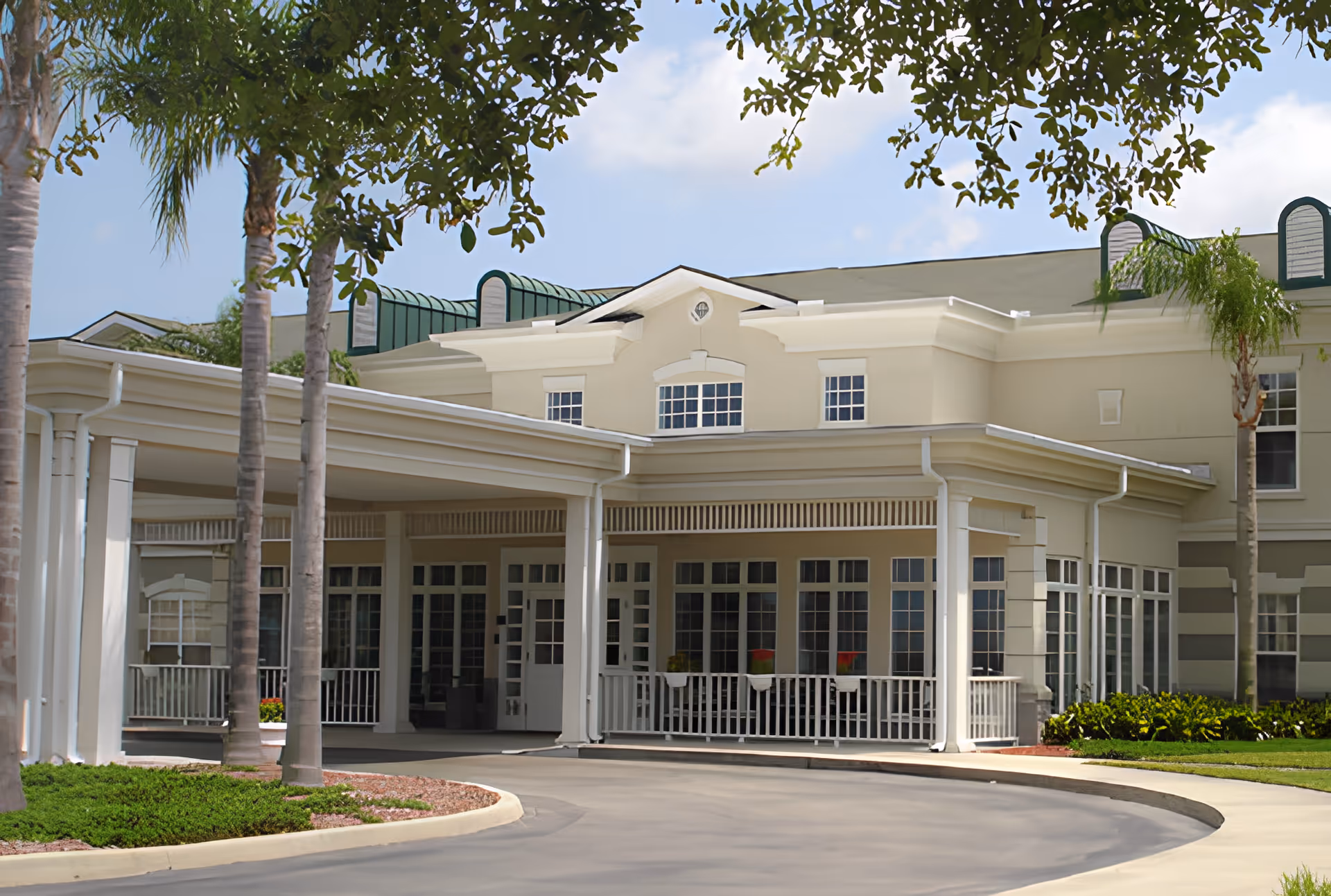 Front entrance of a light-colored senior living facility with a covered porte-cochère, columns, large windows, and palm trees.