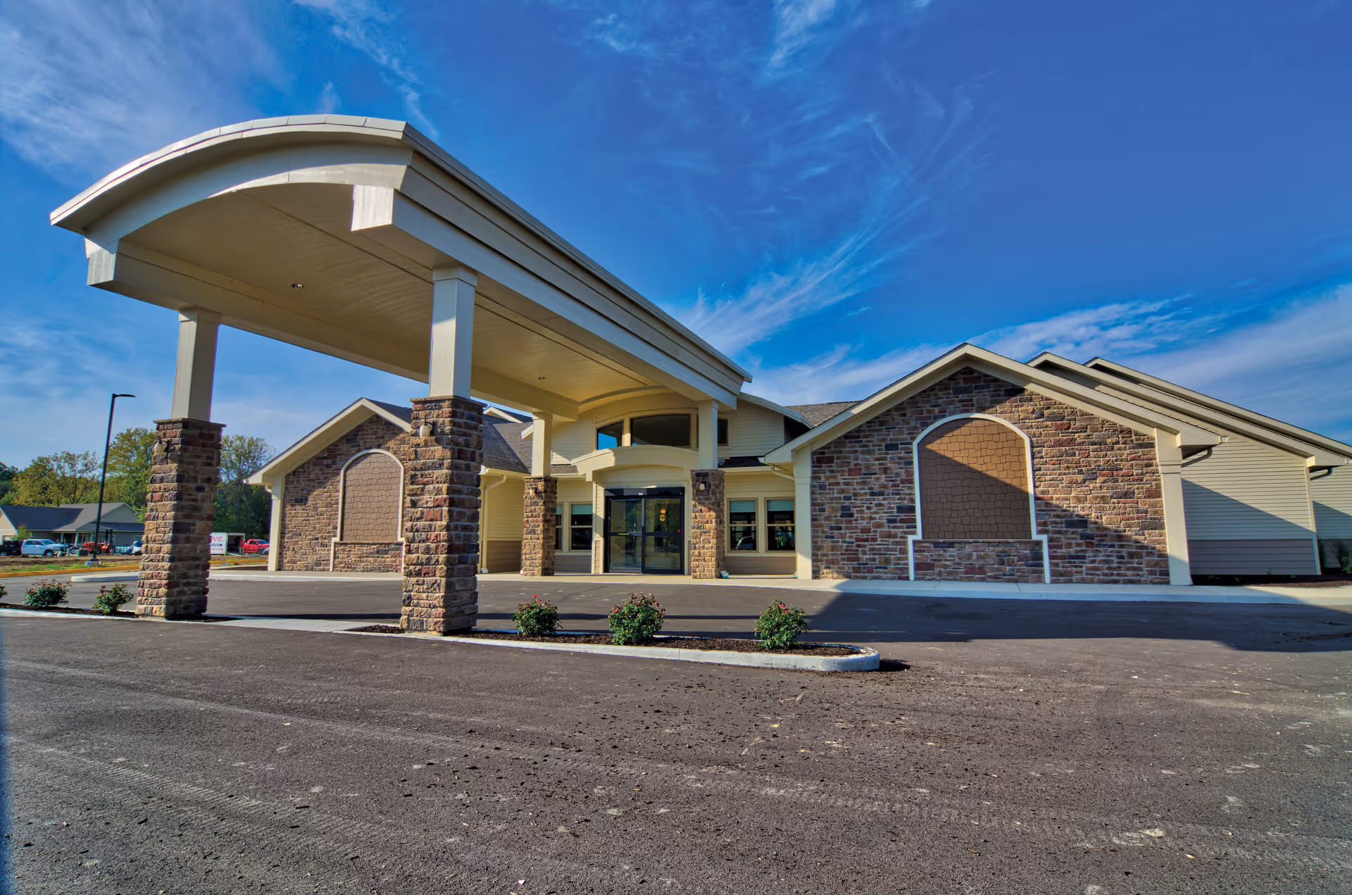 Front exterior of a single-story memory care building with a covered porte-cochere and stone facade under a blue sky.