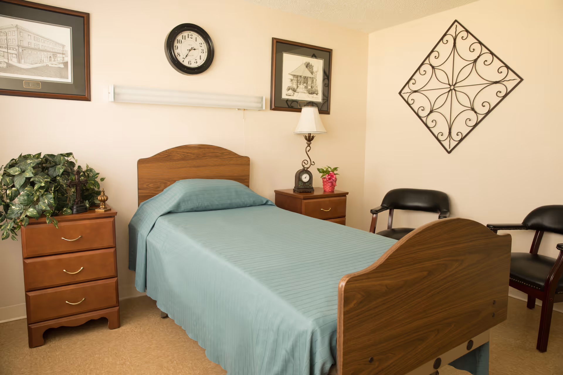 A simple bedroom in a senior living facility with a single wooden bed covered with a teal bedspread. There are two wooden nightstands on either side of the bed, one with a green plant and the other with a lamp and a small clock. Two black chairs are placed against the wall, which is decorated with framed pictures, a clock, and a decorative metal wall hanging.