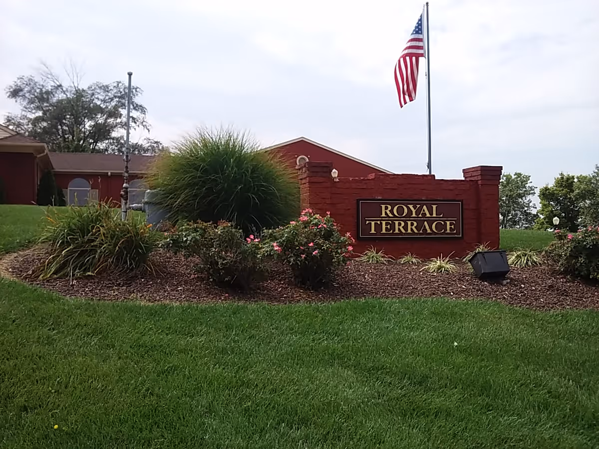 Red brick entrance sign reading Royal Terrace, surrounded by shrubs and an American flag.