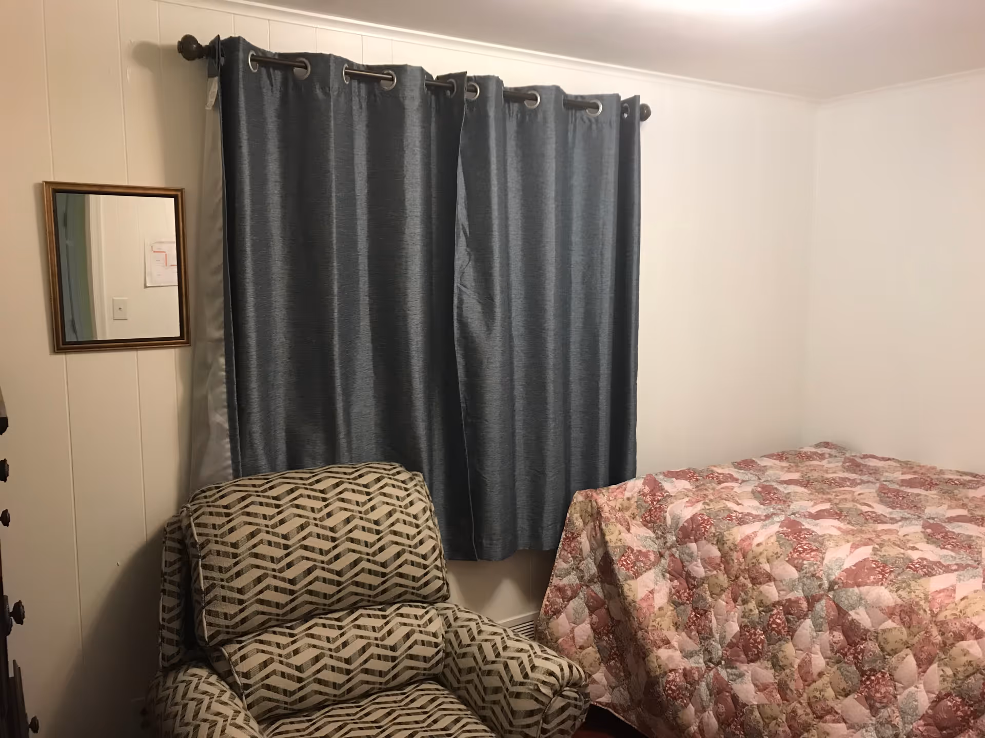 Small bedroom with a patterned armchair, blue curtains, and a floral quilted bed.