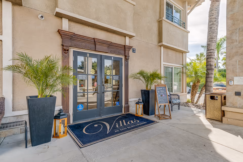 Exterior entrance of Villas at San Bernardino with double doors, potted plants, and a welcome sign.