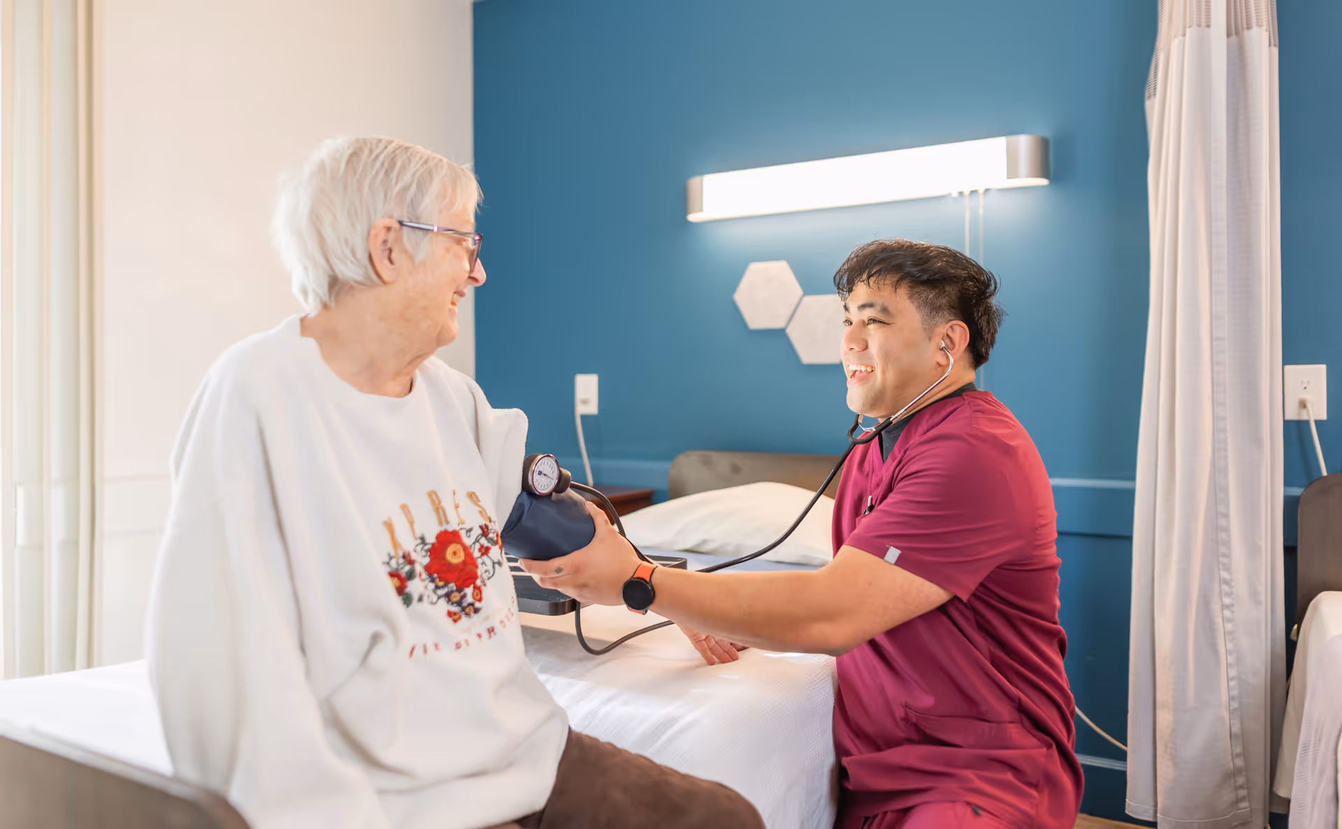 A healthcare worker in maroon scrubs is smiling and measuring the blood pressure of an elderly woman with white hair and glasses, who is sitting on a bed in a room with blue walls and a white curtain.