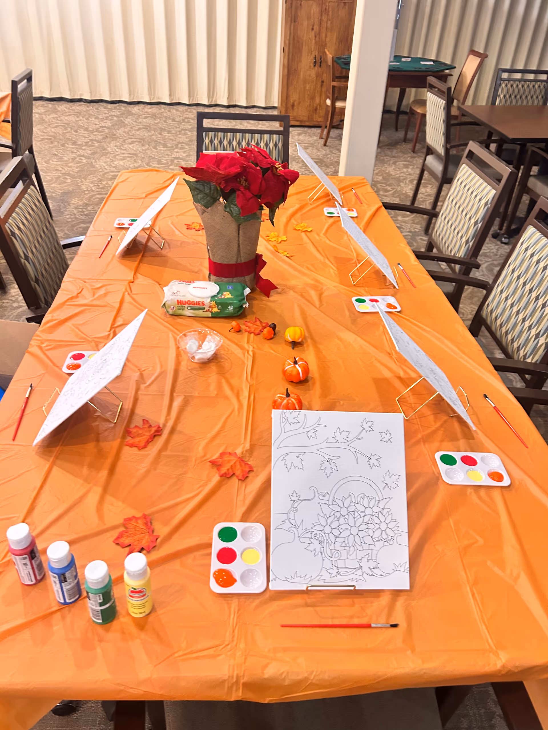 Indoor activity table with painting supplies, orange tablecloth, centerpiece, and autumn decorations.