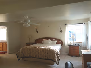 A bedroom with a large bed centered against the wall, two bedside tables with lamps, a ceiling fan, a window with white curtains letting in natural light, and a desk with a chair in the foreground. The room has light-colored walls and carpeted floor.