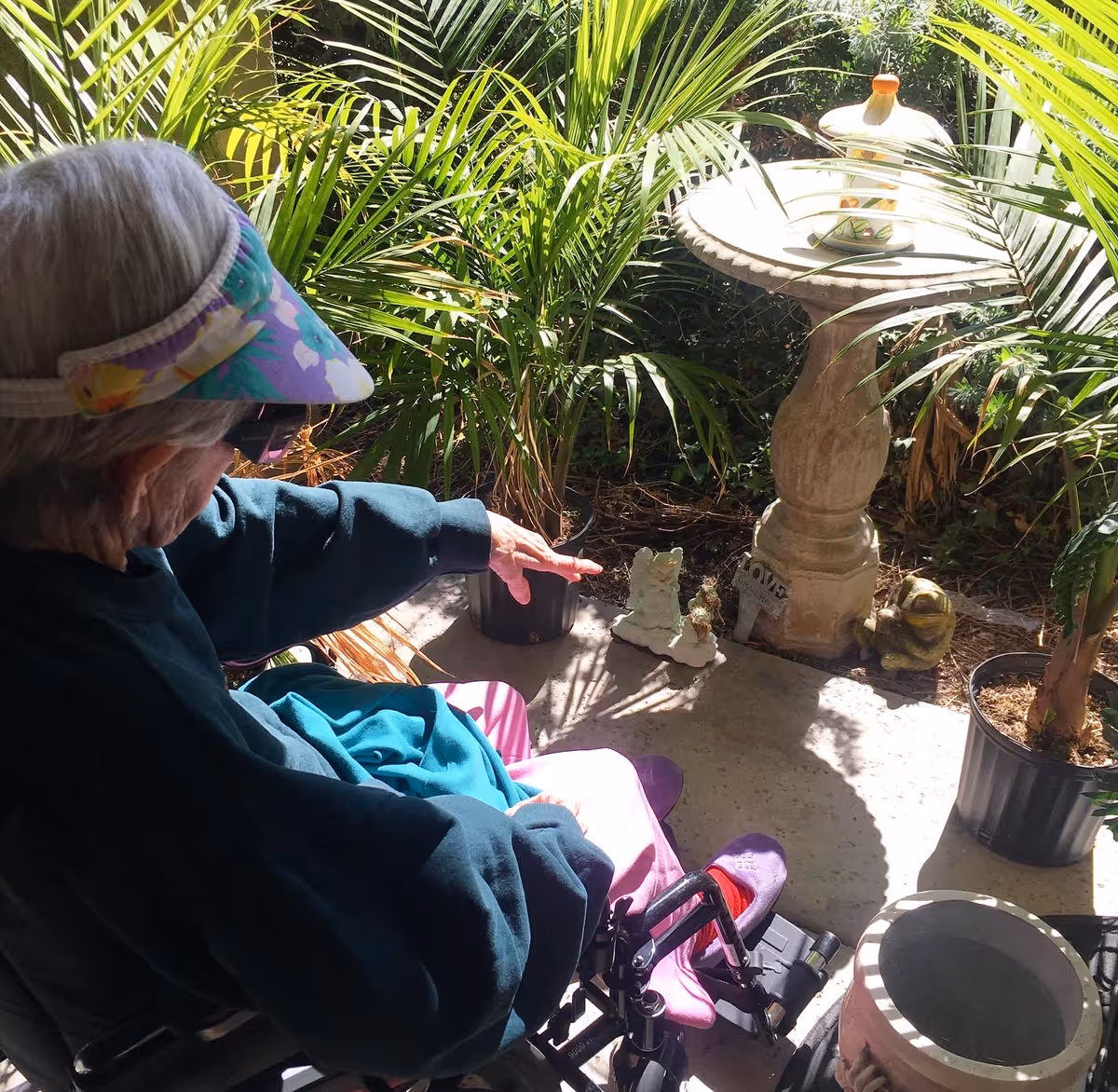 An elderly person wearing a colorful visor and dark clothing sits in a wheelchair outdoors, pointing towards small garden statues near a birdbath surrounded by green plants.