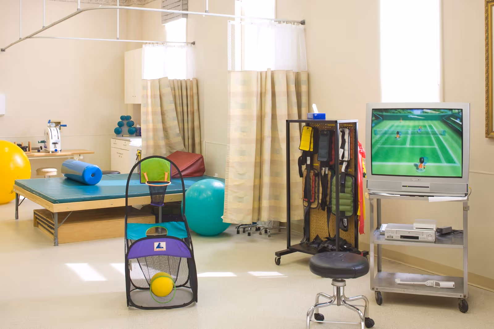 A physical therapy or rehabilitation room with exercise equipment including large exercise balls, a padded therapy table, resistance bands, and a small basketball hoop game. There is a TV on a cart displaying a tennis video game, and privacy curtains hanging from ceiling tracks.