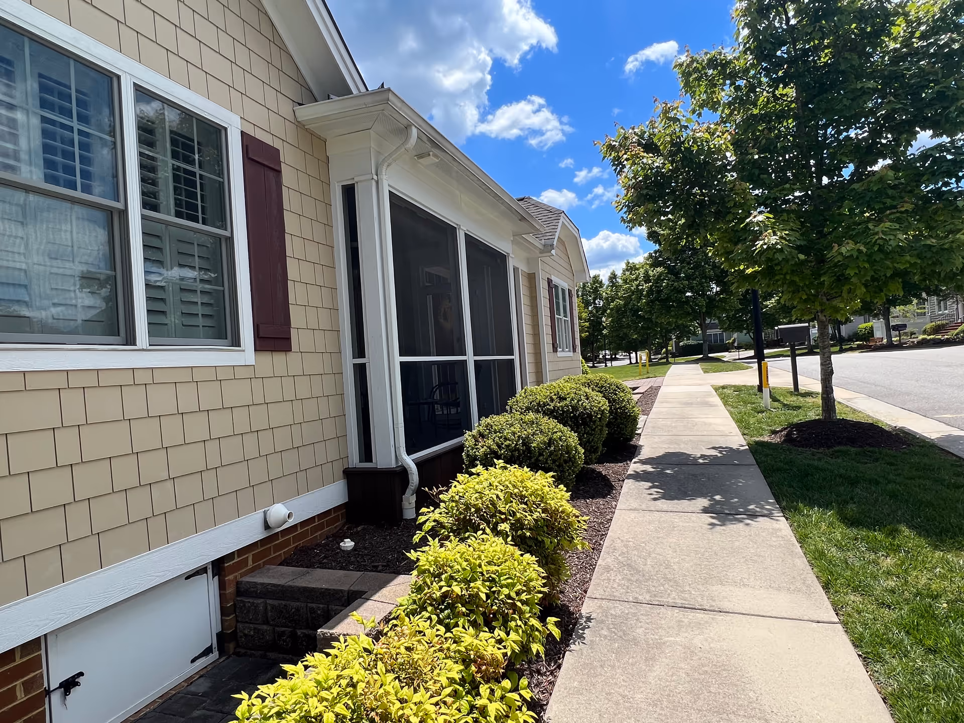 Side view of a beige residential building with maroon shutters and a screened porch. There are neatly trimmed bushes along the building and a sidewalk running parallel to it. Trees line the sidewalk under a partly cloudy blue sky.