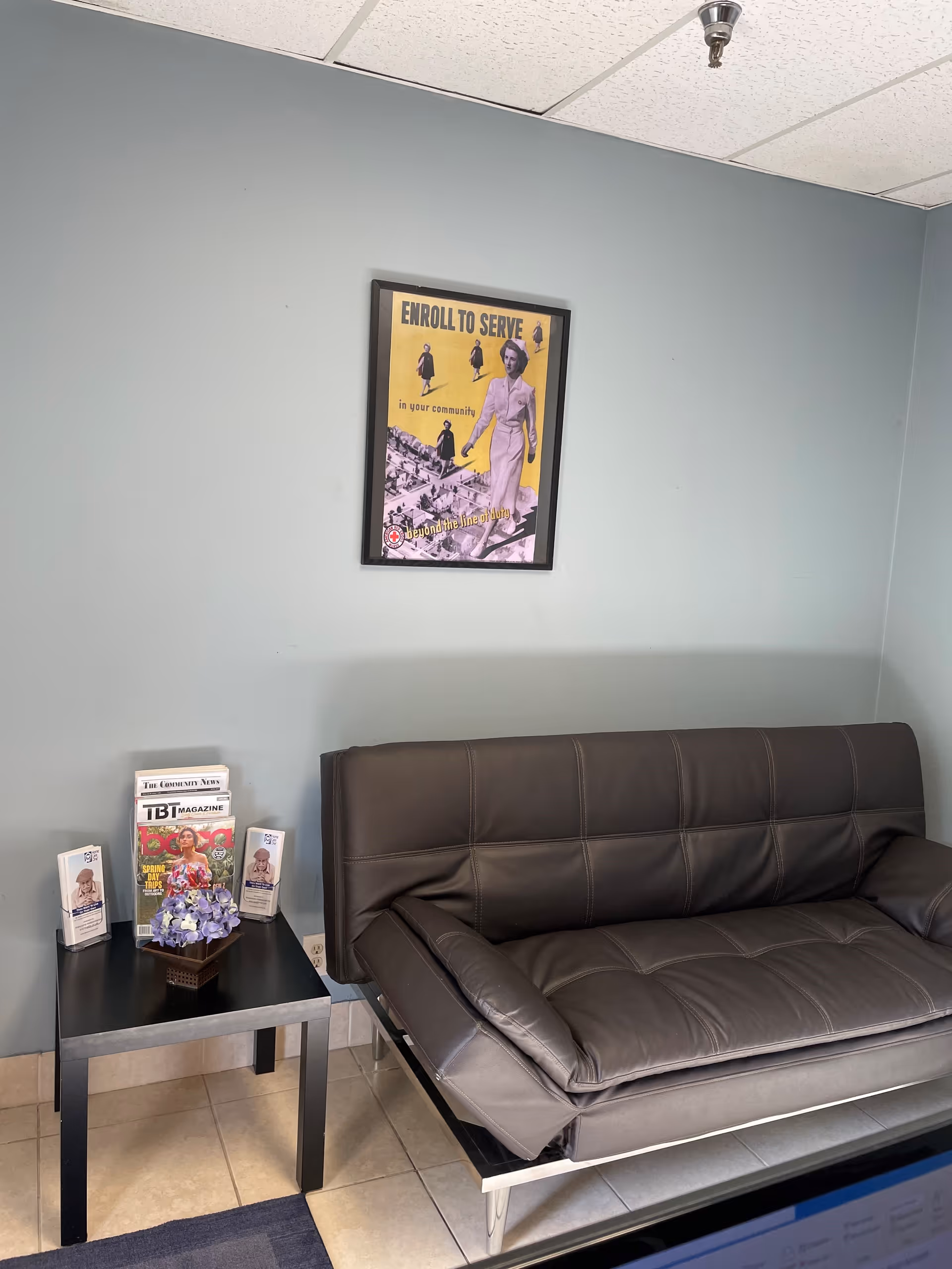 A small waiting area with a gray leather sofa, side table, magazines, brochures, and wall poster.