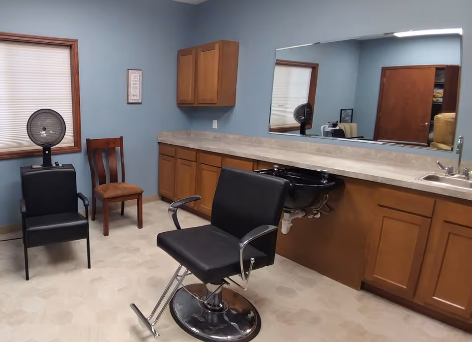 Interior of a senior living facility with a barber chair, sink, mirror, and cabinets.