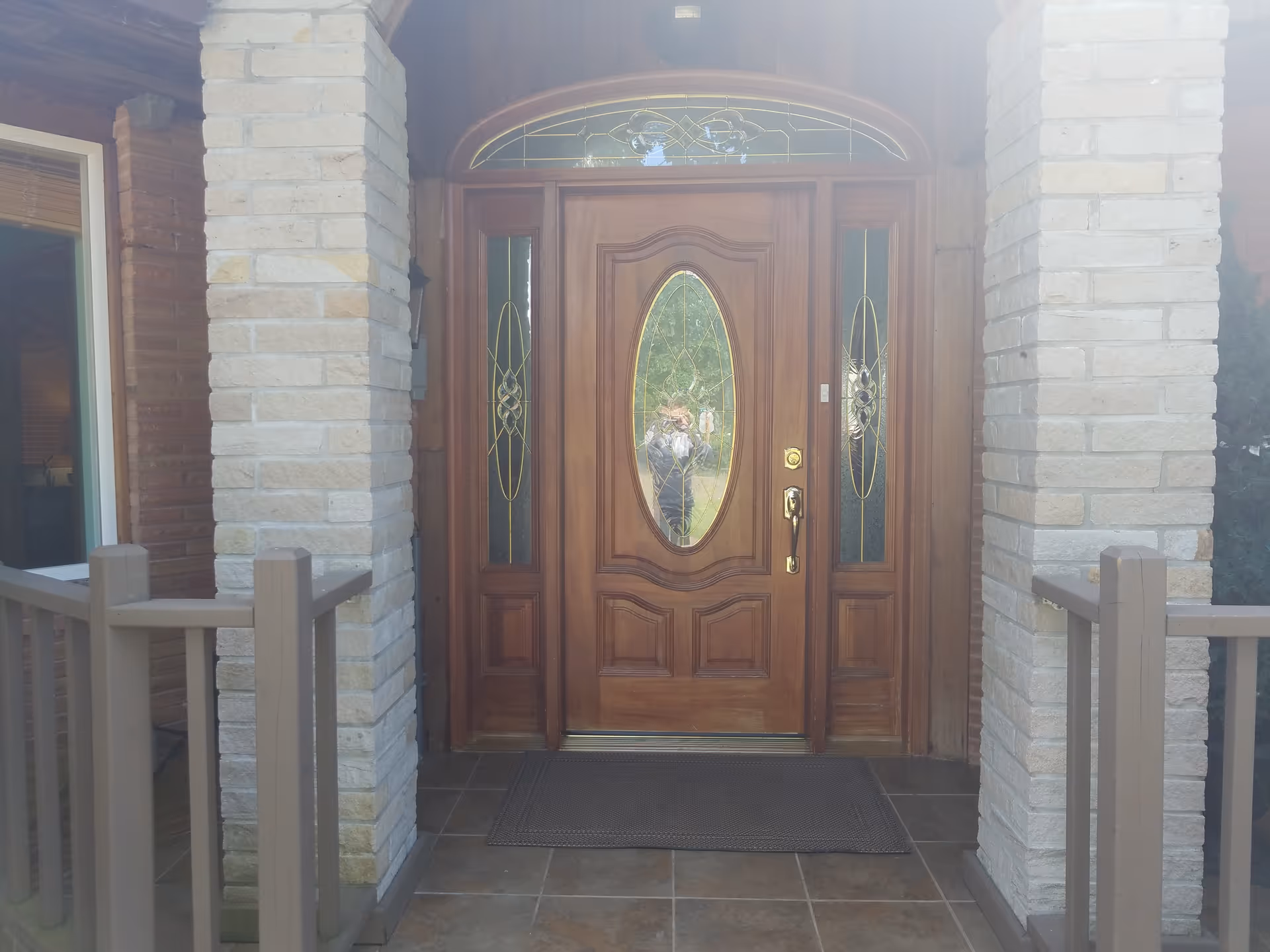Wooden front entrance door with oval glass window flanked by sidelights, stone columns, and railing.