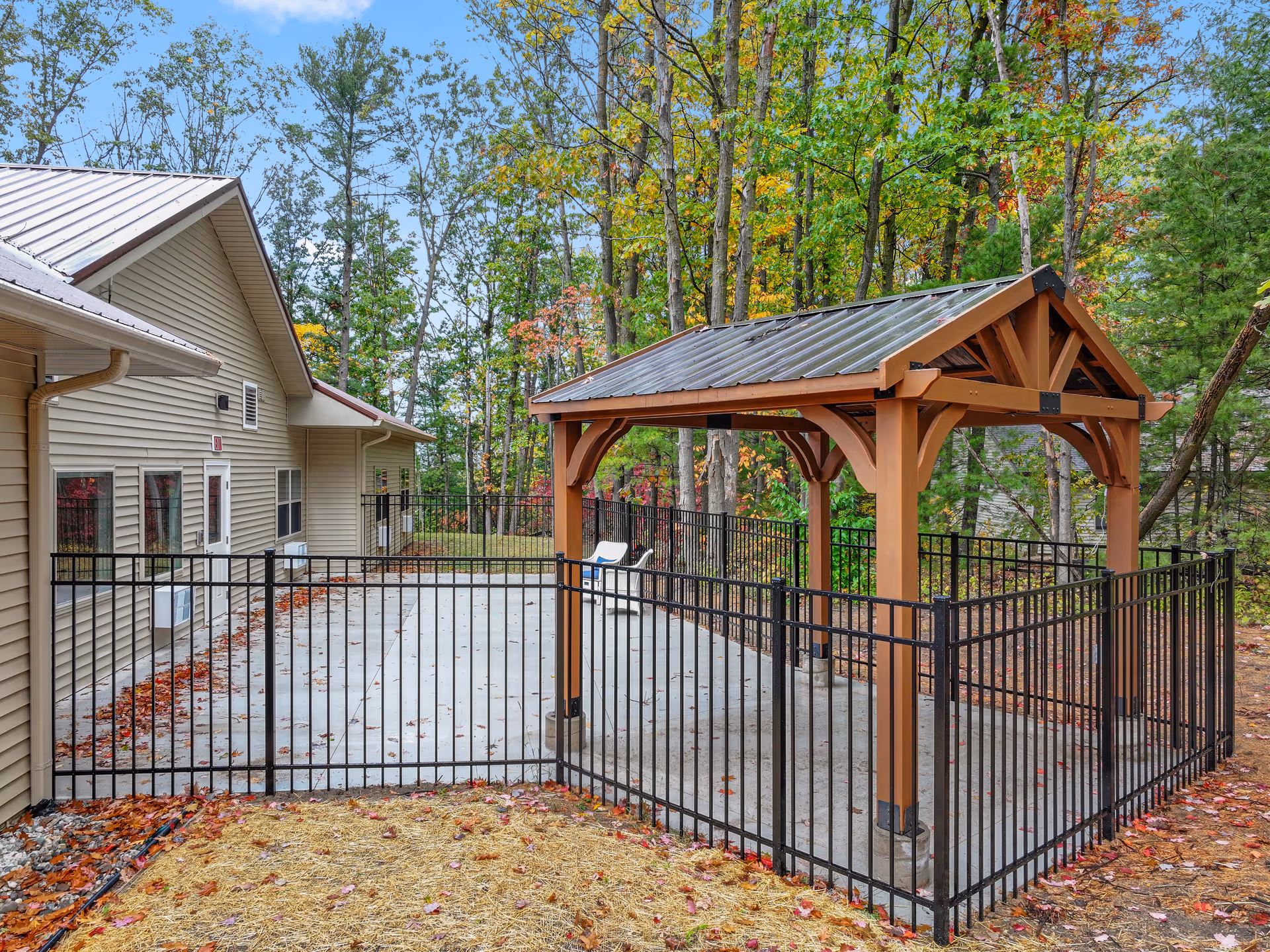 Outdoor fenced patio with a wooden gazebo beside a beige building, surrounded by trees.