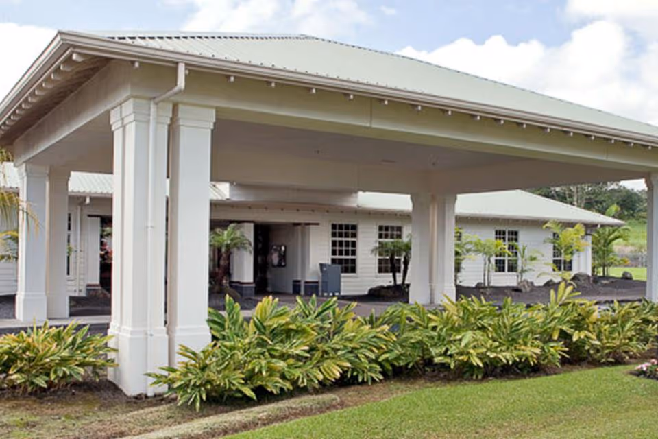 White building with a covered entrance, columns, and lush landscaping in front.