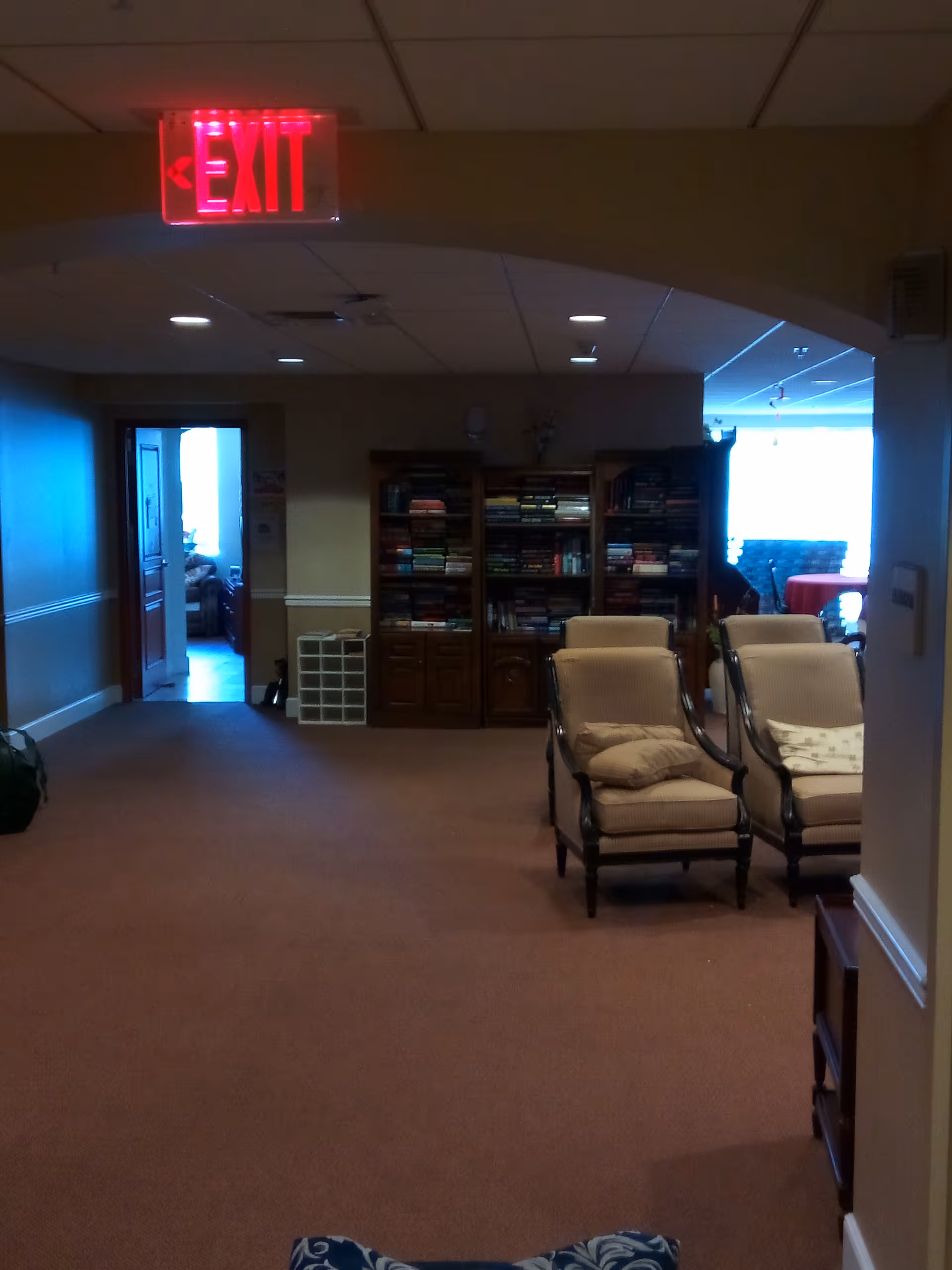 Lounge seating in a corridor with beige chairs, bookshelves, and an illuminated exit sign.