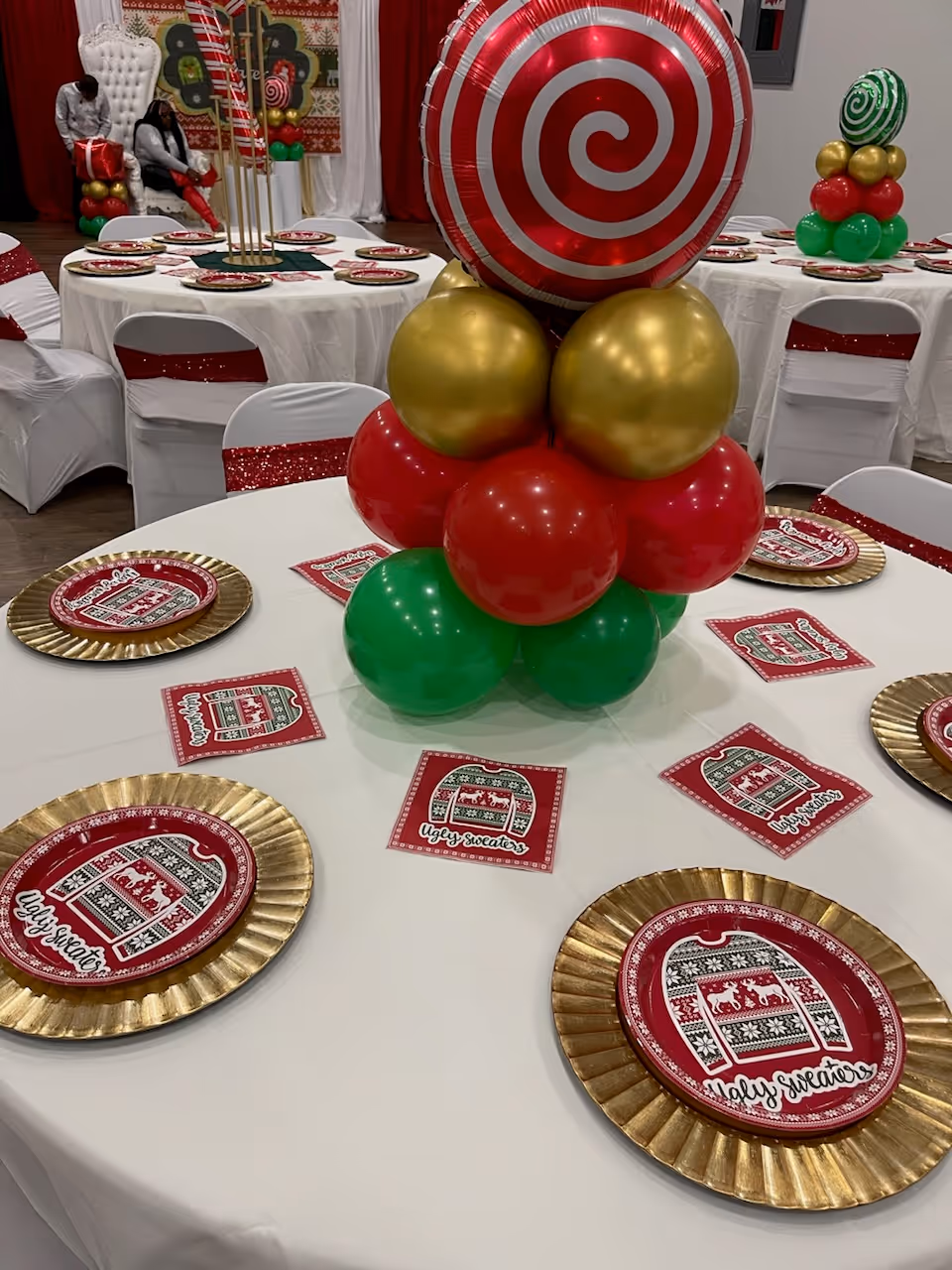 A decorated dining room with round tables, gold charger plates, red napkins, and a balloon centerpiece.