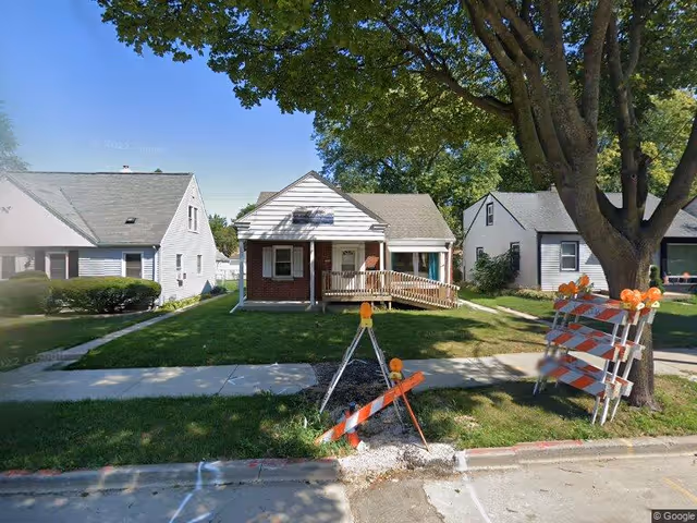 Small single-story house with a front wheelchair ramp, a large tree in the yard, and traffic barricades on the lawn and sidewalk.