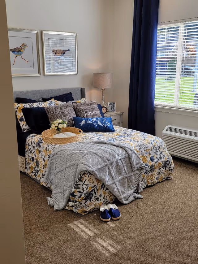 Cozy bedroom with floral duvet, decorative pillows, slippers, and blue curtains.