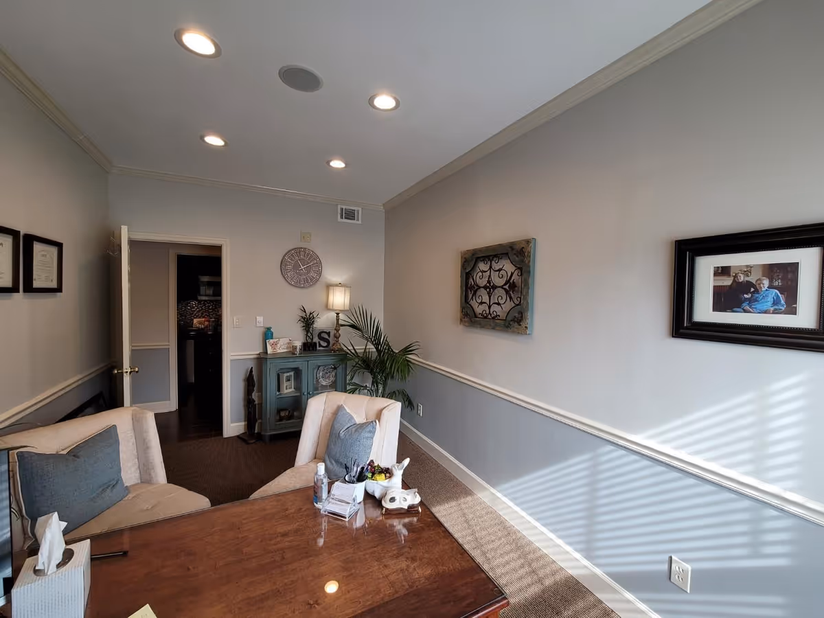 Cozy senior living lounge with a wooden table, chairs, plants, and wall decor.