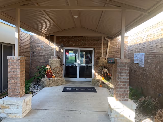 Covered entrance to a brick building with double glass doors, a doormat, brick columns, and hay-bale scarecrow decorations.